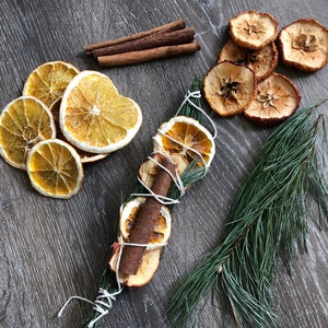 May include: Dried orange slices, apple slices, and cinnamon sticks arranged on a dark wooden surface. A bundle of dried fruit and spices is tied with white twine, alongside a sprig of pine. The arrangement evokes a rustic, natural aesthetic.