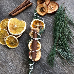 May include: Overhead shot of dried orange and apple slices, cinnamon sticks, and a bundle of dried fruit and cinnamon tied with twine. A sprig of pine needles is also present. The items are arranged on a dark wood-grain surface.