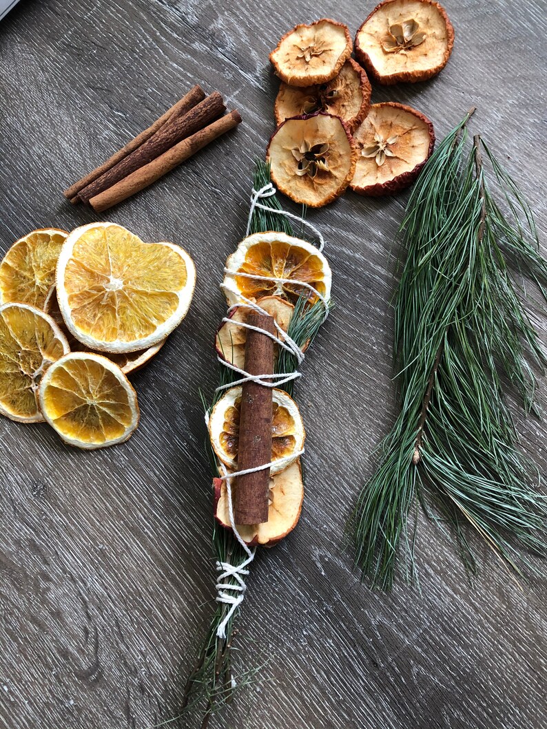 May include: Overhead shot of dried orange and apple slices, cinnamon sticks, and a bundle of dried fruit and cinnamon tied with twine. A sprig of pine needles is also present. The items are arranged on a dark wood-grain surface.