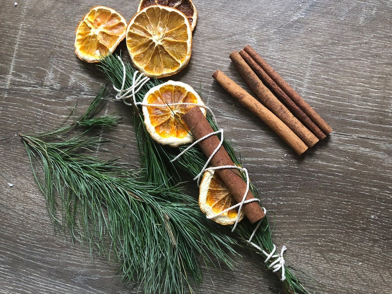 Peut inclure: Composition aromatique avec des tranches d'orange s&eacute;ch&eacute;es, des b&acirc;tons de cannelle et un bouquet d'aiguilles de pin attach&eacute;es avec de la ficelle blanche. L'ensemble est pos&eacute; sur une surface en bois fonc&eacute;, cr&eacute;ant une esth&eacute;tique rustique et naturelle.