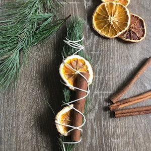 Peut inclure: Un bouquet aromatique d'aiguilles de pin vertes, un b&acirc;ton de cannelle et des tranches d'orange s&eacute;ch&eacute;es, envelopp&eacute; de ficelle blanche. Plusieurs b&acirc;tons de cannelle et tranches d'orange s&eacute;ch&eacute;es sont dispers&eacute;s sur une surface en bois.