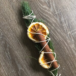 Peut inclure: Un bouquet aromatique compos&eacute; de brins de pin vert, d'un b&acirc;ton de cannelle et de deux tranches d'orange s&eacute;ch&eacute;es. Les ingr&eacute;dients sont li&eacute;s avec de la ficelle blanche, cr&eacute;ant une esth&eacute;tique rustique et naturelle. Id&eacute;al pour la d&eacute;coration ou la parfumerie d'int&eacute;rieur.