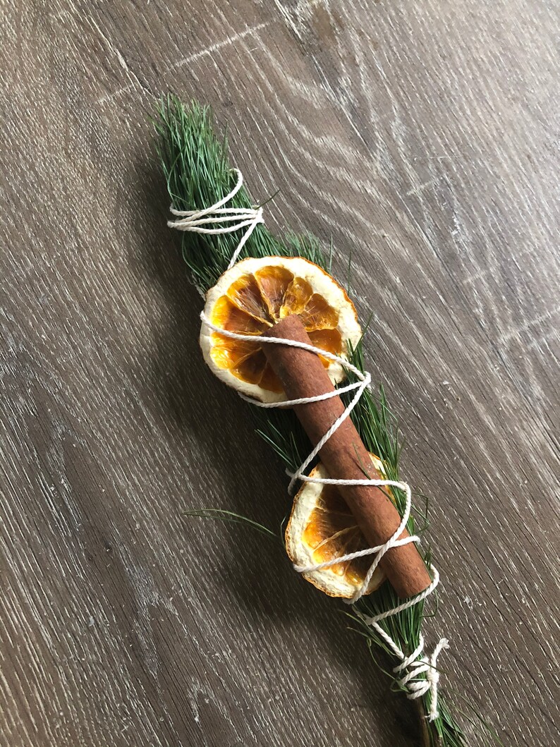 Peut inclure: Un bouquet aromatique compos&eacute; de brins de pin vert, d'un b&acirc;ton de cannelle et de deux tranches d'orange s&eacute;ch&eacute;es. Les ingr&eacute;dients sont li&eacute;s avec de la ficelle blanche, cr&eacute;ant une esth&eacute;tique rustique et naturelle. Id&eacute;al pour la d&eacute;coration ou la parfumerie d'int&eacute;rieur.