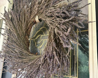May include: A circular wreath constructed from dried lavender stems with a muted purple and grey color palette. The stems are arranged in a clockwise direction, secured to a base, and feature numerous small, dried flower buds throughout.