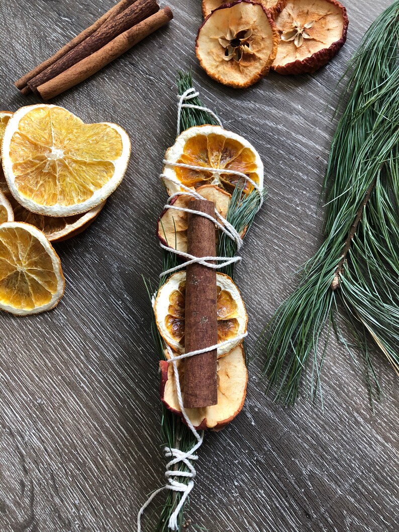 May include: Aromatic arrangement featuring dried orange and apple slices, a cinnamon stick, and evergreen sprigs, bound with twine. Additional cinnamon sticks and dried fruit are scattered around. The composition is set against a wood-grain surface.