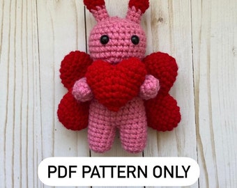 Peut inclure: Un jouet amigurumi en crochet rose avec des ailes rouges et un cœur rouge. Le jouet a des yeux noirs et une expression douce.  MODÈLE PDF UNIQUEMENT.