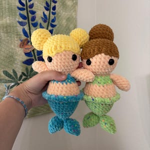 May include: Two handmade crochet mermaid dolls. One doll has yellow hair and a blue tail, the other has brown hair and a green tail. Both dolls have peach-colored skin, black eyes, and small arms.