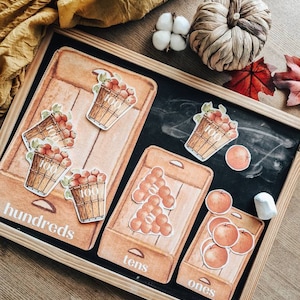May include: A wooden framed chalkboard featuring apple-themed educational resources. The board shows the words "hundreds", "tens", and "ones" with apple illustrations. Decorative items include a woven pumpkin, cotton balls, and autumn leaves.