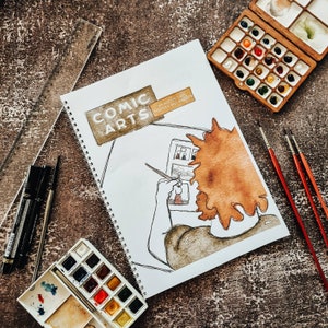May include: A comic arts sketchbook with a spiral binding. The cover features a drawing of a person with brown hair, holding a paintbrush and looking at a page of sketches. The text on the cover reads "Comic Arts: An Art Journal for Theories and Experiments."
