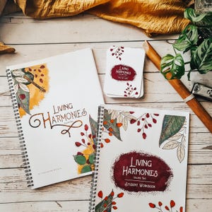 Op de afbeelding: Twee spiraalgebonden werkboeken met de titel "Living Harmonies" op de omslag. De werkboeken liggen op een houten oppervlak met een bruine stof en een groene plant op de achtergrond. Het werkboek links is deel één en het werkboek rechts is deel twee, een werkboek voor studenten.