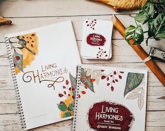 Peut inclure: Deux cahiers à spirales avec le titre "Living Harmonies" sur la couverture. Les cahiers sont sur une surface en bois avec un tissu brun et une plante verte en arrière-plan. Le cahier de gauche est le volume un et le cahier de droite est le volume deux, un cahier d'exercices pour les élèves.