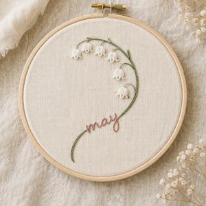 May include: Embroidered hoop art with a lily of the valley design. The artwork features white flowers, green stems, and the word "may" in a cursive font. The embroidery is set in a wooden hoop.