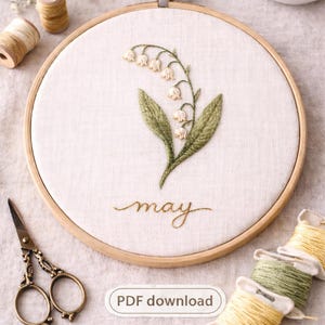 Op de afbeelding: Geborduurde borduurringkunst met een lelietje-van-dalen ontwerp met groene bladeren en witte klokvormige bloemen. Het woord "may" is onder het bloemmotief gestikt. Inclusief een houten borduurring, schaar en garenklosjes. Tekst: PDF download.