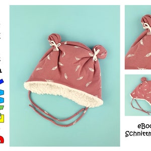 Könnte beinhalten: Eine rosa Strickmütze mit weißem Federmuster und weißen Bommeln oben. Die Mütze hat ein weißes Fleecefutter und eine Bindekordel.