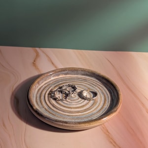 Puede incluir: Un plato de cerámica para anillos con un diseño en espiral en tonos grises y azules contiene tres anillos de plata. El plato se encuentra sobre una superficie con vetas de mármol rosa y blanco, con un fondo verde.