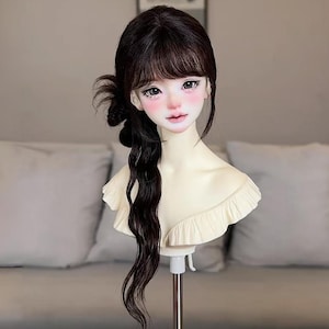 Può includere: Una testa di bambola con lunghi capelli scuri acconciati con frangia e una coda di cavallo laterale. La bambola ha un incarnato pallido con fard rosa e lineamenti dettagliati. La testa è montata su un supporto metallico con una base circolare trasparente e un top a balze color crema.