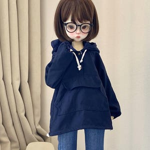May include: A doll wearing a navy blue hooded jacket, blue jeans, and glasses. The doll has brown hair and is wearing sneakers. The jacket has a front pocket and a drawstring hood. The jeans are flared at the bottom.