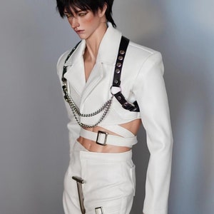 May include: A white cropped jacket with a black leather harness and silver chain. The jacket has a silver buckle and a white belt. The model is wearing white trousers with silver buckles.
