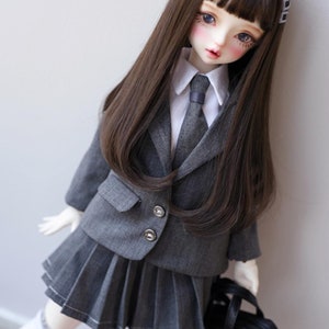 Puede incluir: Una muñeca vestida con un uniforme escolar gris, que incluye una chaqueta, una falda plisada y una corbata. La muñeca tiene el pelo largo castaño y lleva un bolso negro. El uniforme presenta botones plateados y detalles blancos.
