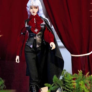 May include: A doll dressed in a gothic style. The doll is wearing a red blouse, black vest, and trousers. A black cape and boots complete the look. The doll has white hair and is positioned in front of a red curtain.