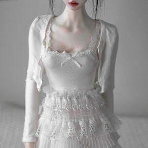 Op de afbeelding: Een witte pop die een witte kanten jurk draagt met een wit vestje. De pop heeft rode lippen en zwart haar.