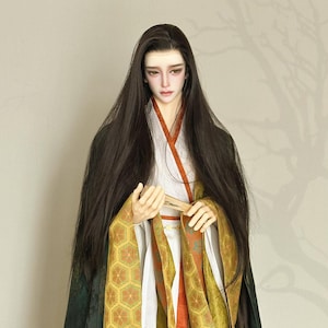 Puede incluir: Una muñeca vestida con una túnica japonesa tradicional con intrincados patrones dorados y verdes. La túnica tiene mangas largas y fluidas y está atada a la cintura con una faja. La muñeca tiene el pelo largo y negro y sostiene un abanico.