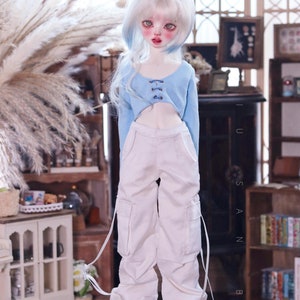 May include: A doll wearing a light blue crop top with a lace-up detail and white cargo pants with adjustable straps. The doll has long, white hair with blue highlights and is wearing black shoes.