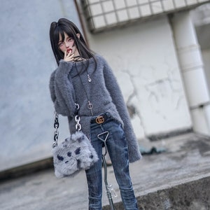 May include: A doll in a grey fluffy jumper, blue jeans, and black boots with gold details. The doll carries a small grey bag with a chain strap and a black belt with a gold buckle. The doll is standing on a concrete surface.
