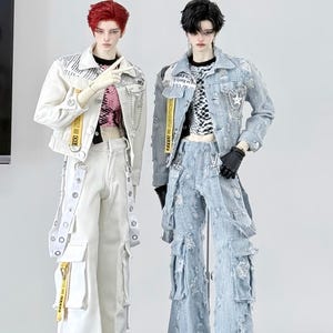May include: Two fashion dolls wearing stylish outfits. One doll is in a white jacket and pants with yellow accents, the other in a light blue denim jacket and distressed jeans. Both dolls have unique hairstyles and are posed in a studio setting.