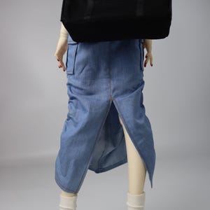 May include: A doll wearing a denim skirt with a front slit, white socks, and white sneakers with black accents. A black duffel bag is slung over the doll's shoulder. The doll's nails are painted red.