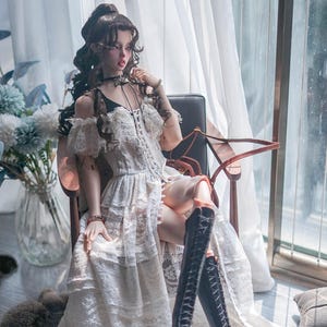May include: A white lace dress with a sweetheart neckline and off-the-shoulder straps. The dress has a tiered skirt and is worn with black knee-high boots.