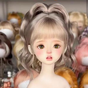 Puede incluir: Una cabeza de muñeca con piel pálida, grandes ojos color avellana y mejillas sonrosadas. La muñeca tiene cabello castaño claro ondulado peinado en una coleta alta con flequillo que enmarca el rostro y una expresión con labios entreabiertos.