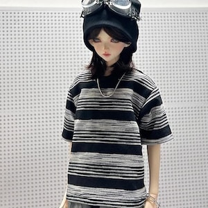 Pode incluir: Uma boneca vestindo uma camisa listrada preta e branca, um gorro preto com óculos e calças cinzas.