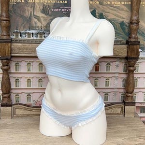Op de afbeelding: Een lichtblauw en wit gestreepte lingerie set op een mannequin. De set bestaat uit een crop top met dunne bandjes en ruches, en een bijpassende lage broek. De achtergrond toont een decoratieve houten structuur en een vintage poster.
