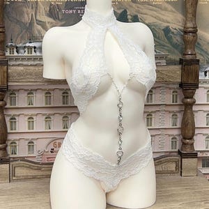 May include: White lace lingerie set on a mannequin. The set includes a halter top and a low-rise bottom, connected by a silver chain. The background features a vintage movie poster with the text "THE BUDAPEST HOTEL".