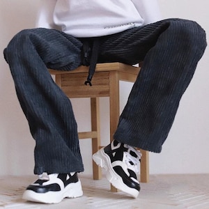 Pode incluir: Uma pessoa vestindo calças de veludo cotelê pretas com uma camisa branca que diz "Essentials". Ela está usando tênis preto e branco com sola branca grossa.