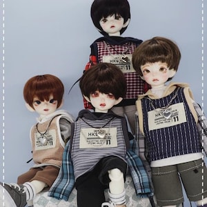 May include: Four dolls wearing different outfits. Each doll has short brown or black hair and is wearing a necklace with a heart pendant. The dolls are wearing a variety of clothing, including plaid shirts, striped shirts, and denim shorts. The dolls are standing on a chequered surface.