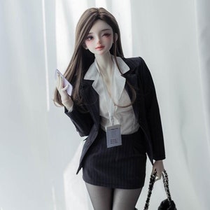 May include: A doll wearing a black blazer, white shirt, and black skirt with a black handbag. The doll is standing on a black stand and has long brown hair.