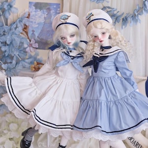 Op de afbeelding: Twee porseleinen poppen in matrozenoutfits. Eén pop draagt een witte jurk met marineblauwe strepen en een bijpassende hoed. De andere pop draagt een lichtblauwe jurk. Beide poppen hebben blond haar en poseren samen.