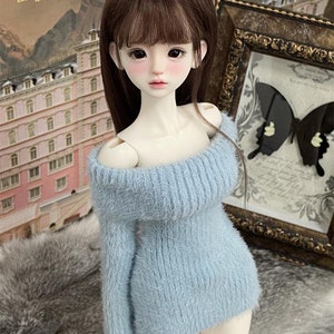 Puede incluir: Un vestido de punto azul claro y peludo con hombros descubiertos en una mu&ntilde;eca con cabello largo y casta&ntilde;o. La mu&ntilde;eca tiene piel clara y rubor rosado en las mejillas.