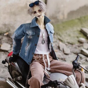 Puede incluir: Una muñeca con una chaqueta vaquera azul, un top blanco y pantalones de pana marrón está sentada en una motocicleta plateada. La muñeca tiene el pelo plateado y lleva gafas de sol.