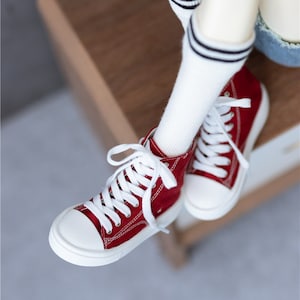 May include: Red high-top canvas sneakers with white laces and white soles. The sneakers are worn by a doll with white socks and black stripes.
