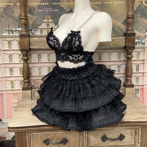 Op de afbeelding: Een zwarte kanten bh en rok set met een parelketting op een mannequin. De mannequin staat op een houten tafel met een lade. De achtergrond is een poster met de tekst "The Grand Budapest Hotel" en een lijst met acteurs.