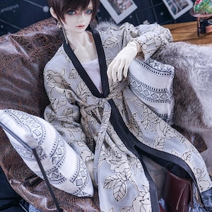 May include: A doll wearing a light gray kimono-style robe with a black trim and a floral pattern. The doll is seated with a matching pillow. The doll has brown hair and is wearing brown boots.