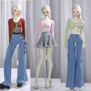 May include: Three dolls wearing different colored crop tops with a button detail and blue denim pants with lace-up details. The dolls are standing on white stands.