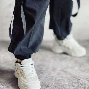 Puede incluir: Zapatillas blancas con detalles negros y un panel de malla. Las zapatillas tienen una suela gruesa y se llevan con pantalones cargo negros.