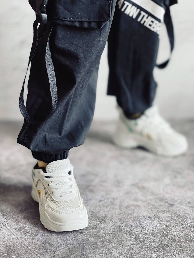 Puede incluir: Zapatillas blancas con detalles negros y un panel de malla. Las zapatillas tienen una suela gruesa y se llevan con pantalones cargo negros.