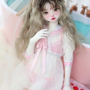 May include: A white doll with long, wavy, light brown and blonde hair wearing a pink and white sailor-style dress with a pink ribbon. The doll is wearing a pink cat ear headband and is sitting in a pink and white inflatable ring with strawberries.
