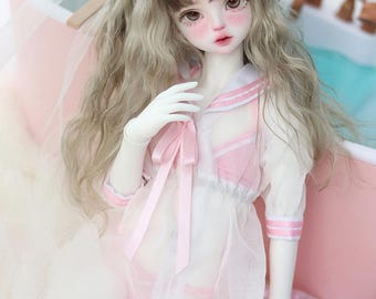 May include: A white doll with long, wavy, light brown and blonde hair wearing a pink and white sailor-style dress with a pink ribbon. The doll is wearing a pink cat ear headband and is sitting in a pink and white inflatable ring with strawberries.
