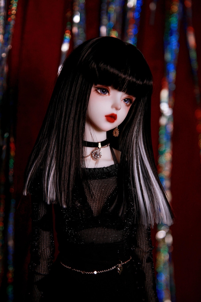 May include: A black and white doll with long hair, wearing a black mesh top, a black choker necklace with a crystal pendant, and a gold chain belt. The doll has blue eyes and red lips.